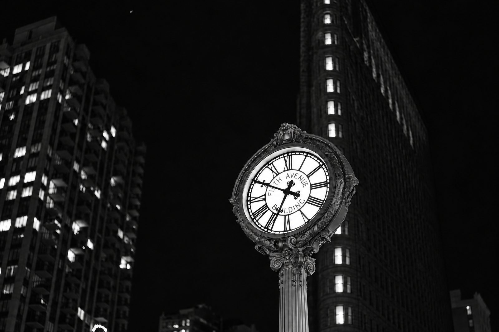 A cast-iron street clock lit up on a New York sidewalk at night