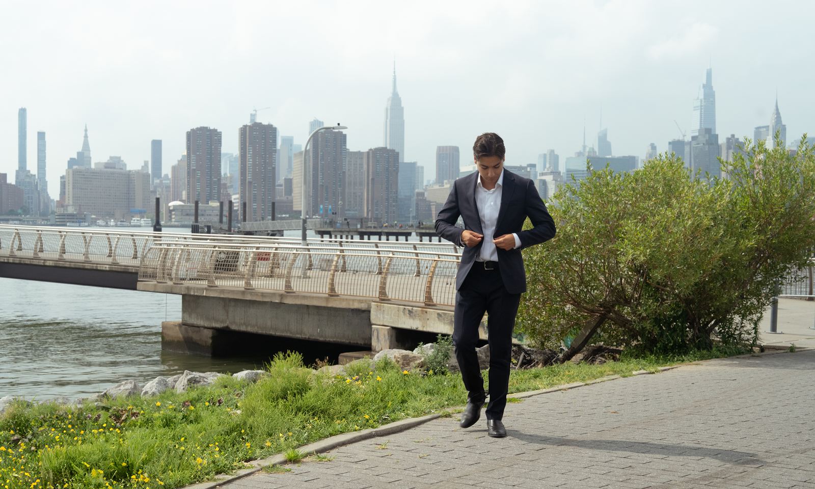 Glenn Velasco walking along a waterfront path with the Manhattan skyline and the Empire State Building behind him